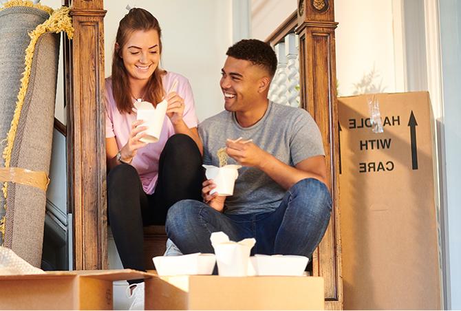 A couple eating together surrounded by moving boxes.